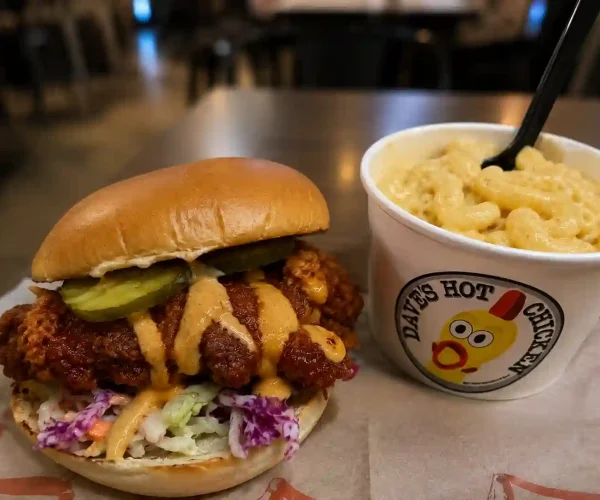 Dave’s Hot Chicken Secret Menu sandwich with crispy chicken, pickles, sauce, and mac and cheese on a restaurant table.